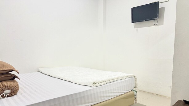 A single bed with a white mattress and a beige headboard, a small bedside table, and a wall-mounted TV. - Cozy Solo Room at Medina House (Banda Aceh)