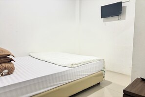 Un letto singolo con materasso bianco e testiera beige, un comodino e una TV fissata al muro.