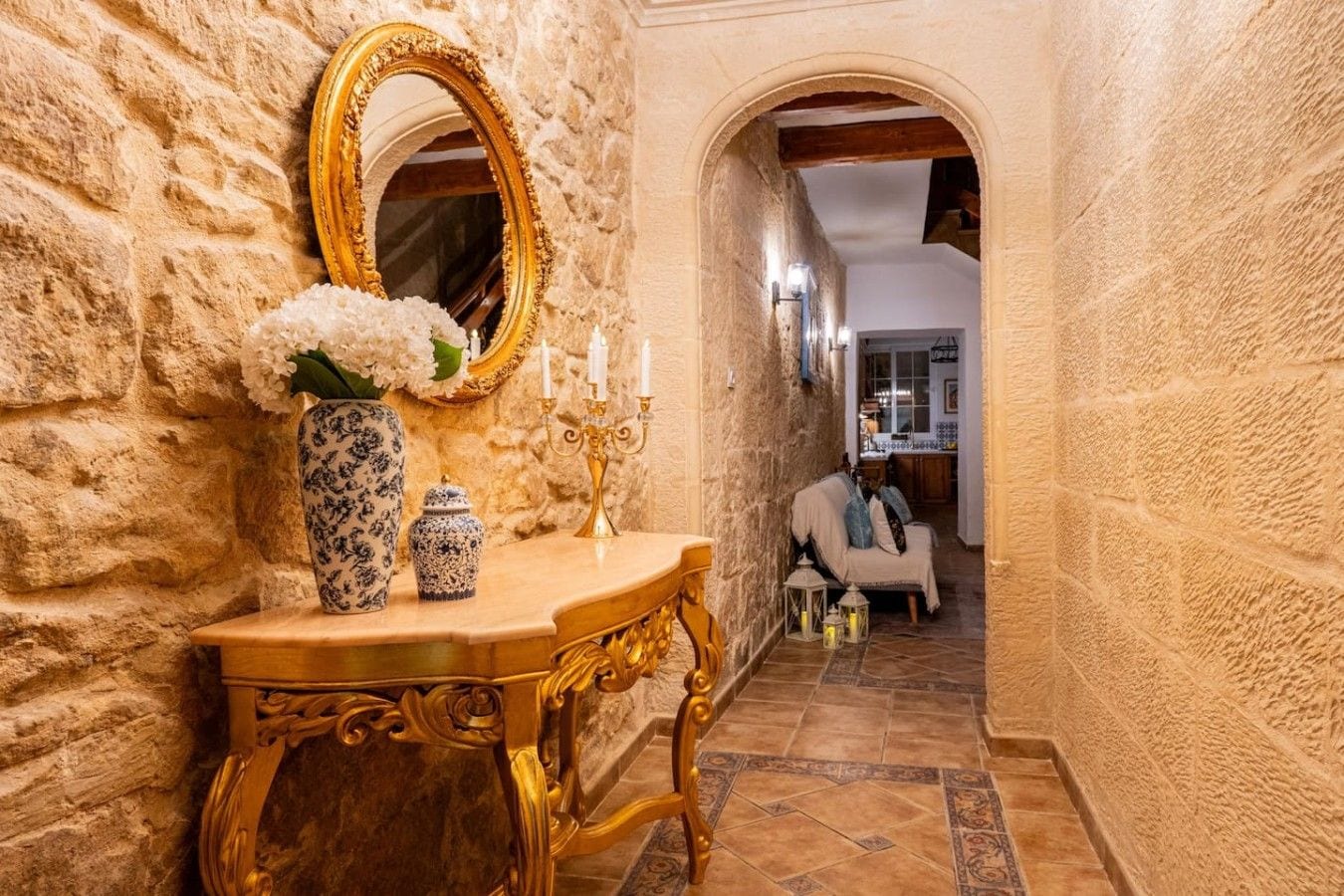 A hallway with a stone wall, a gold-framed mirror, a vase with flowers, a candelabra, and a sofa in the background.