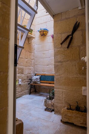 Un cortile stretto con pareti in pietra, una panchina, un punto luce a parete e una coppia di forbici appese al muro.