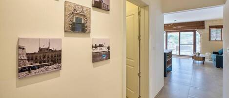 A hallway with a series of framed photographs on the wall, a door leading to a room, and a view into a living area with a sofa and a window.