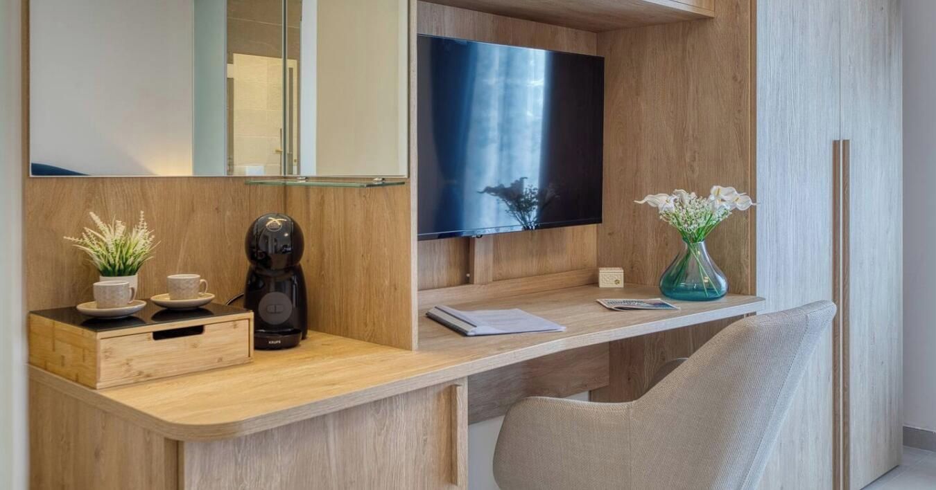 A modern hotel room with a wooden desk, a flat-screen TV, a coffee maker, a vase with flowers, and a comfortable chair.