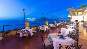 An outdoor dining area with tables and chairs, overlooking a body of water and a cruise ship in the distance. - Beachfront Cabo Escape at Villa del Arco with Pools, Spa & Walkable Location (Cabo San Lucas)