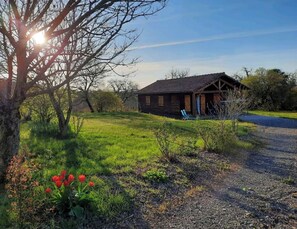 A wooden cabin with a gravel driveway, surrounded by grass and trees.