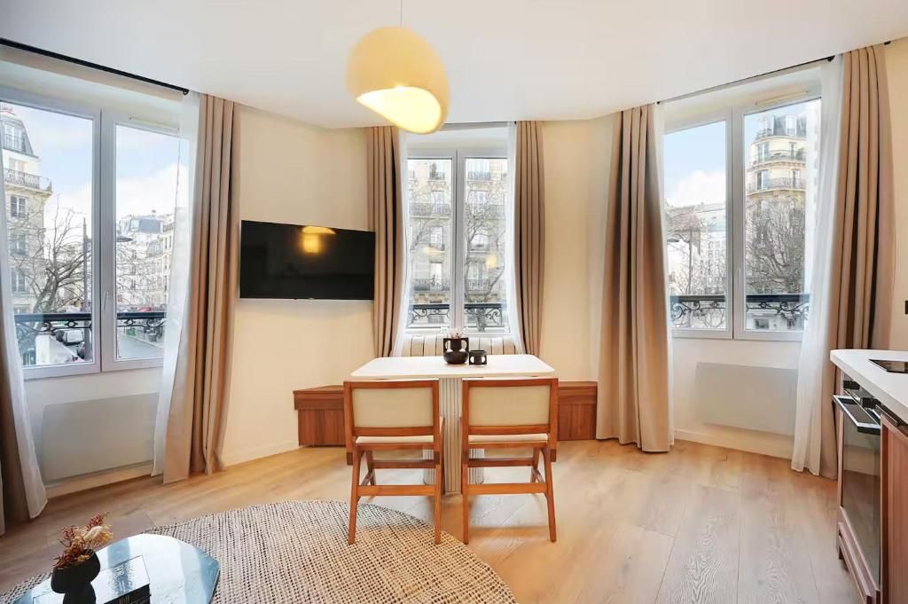 A modern living room with a dining table, two chairs, a flat-screen TV, and large windows with curtains.