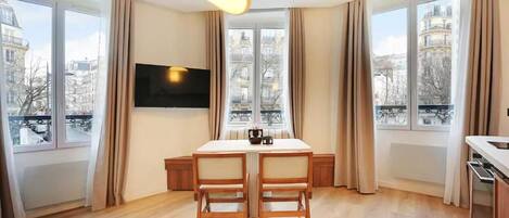 Una sala de estar moderna con una mesa de comedor, dos sillas, un televisor de pantalla plana y ventanas grandes con cortinas.