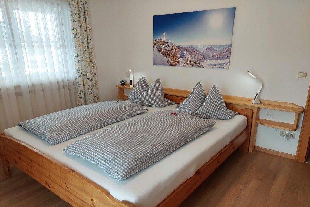 A double bed with checkered bedding, a bedside table with a lamp, a painting on the wall, and a window with curtains.
