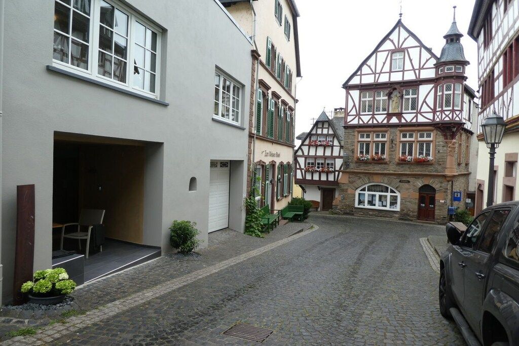 A cobblestone street with traditional half-timbered houses, a car parked on the right, and a building with a visible entrance.