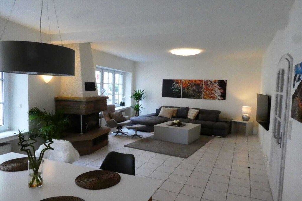 Ein modernes Wohnzimmer mit einer Couch, einem Couchtisch und Sesseln. Ein Kamin, ein Esstisch mit Unterlagen und ein großes Fenster sind ebenfalls vorhanden.