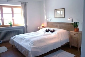Ein Schlafzimmer mit einem Bett, Nachttischen, einem Heizkörper, einem Fenster mit Vorhängen und einer Wandlampe.