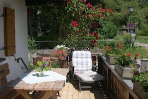 Eine Terrasse im Freien mit einem Holztisch und einem gepolsterten Stuhl, umgeben von Blumentöpfen und blühenden Blumen.