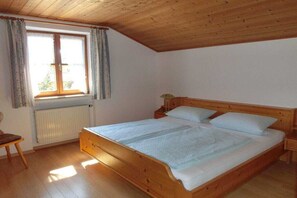 Un dormitori amb un llit de fusta, una tauleta de nit, una finestra amb cortines i un radiador.