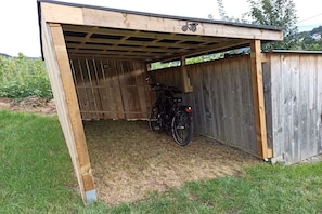 Una caseta de fusta amb una bicicleta a dins, una zona enjardinada a l'exterior i una tanca de fusta al fons.