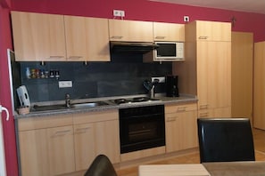 A compact kitchen with a microwave, stove, and oven. There is a sink with a faucet, and cabinets above and below the countertop. A chair is visible in the foreground. - Holiday apartment with 2 bedrooms (Graach)