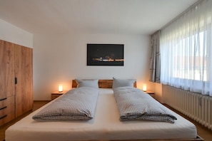 Een tweepersoonsbed met geruit beddengoed, twee nachtkastjes met lampen, een houten garderobe, uitzicht op een nachthemel en dunne gordijnen.