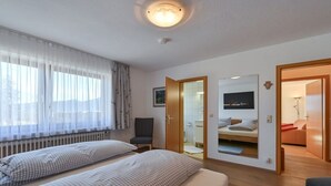 Ein Schlafzimmer mit zwei Betten, einem Fenster mit durchsichtigen Vorhängen, einem Heizkörper, einem Stuhl, einem Spiegel und einer Tür zu einem Badezimmer.