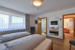 Quarto com duas camas, janela com cortinas leves, radiador, cadeira, espelho e porta que dá para um banheiro.
