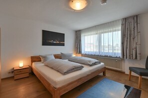 A bedroom with a bed, bedside tables, a nightstand, a chair, a window with curtains, and a wall-mounted picture. - Holiday apartment with 1 bedroom (Füssen)