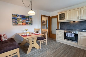 A compact kitchen with a dining area, wooden cabinets, a built-in oven, and a wall-mounted picture. - Holiday apartment with 1 bedroom (Übersee)
