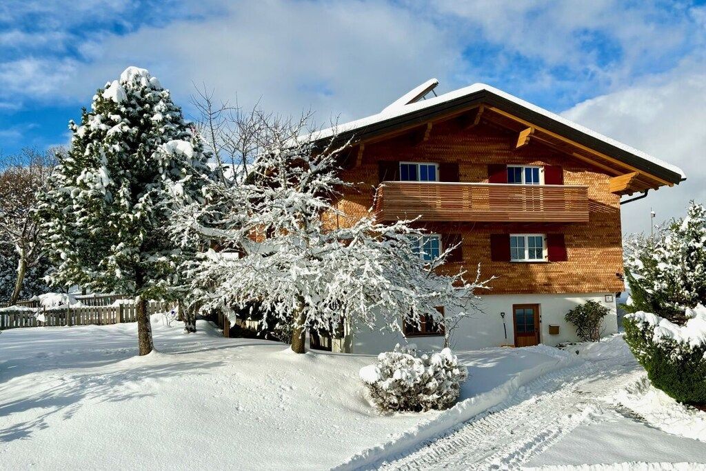 Ein zweigeschossiges Holzhaus mit geneigtem Dach, umgeben von schneebedeckten Bäumen und blauem Himmel.