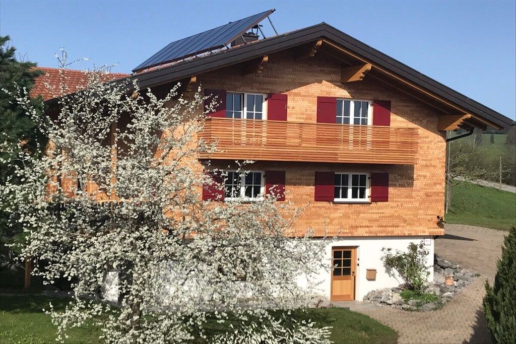 A two-story wooden house with a sloped roof, solar panels, and a red door.
