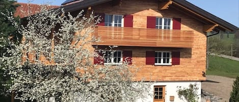 A two-story wooden house with a sloped roof, solar panels, and a red door.