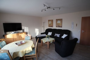 Een woonkamer met een flatscreen televisie, een bank, een eettafel met stoelen en een plafondlamp.
