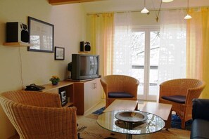 Una sala de estar con muebles de mimbre, una mesa de centro de vidrio, una televisión y vistas a través de una ventana con cortinas.