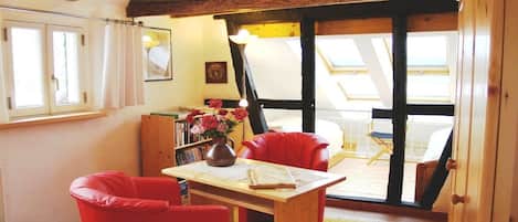 Quarto aconchegante com teto de madeira, uma mesa com um vaso de flores, cadeiras vermelhas e vista através de uma grande janela.