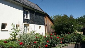 A two-story house with a white facade and dark roof, surrounded by a garden with red flowers. - Holiday apartment with 1 bedroom (Gummersbach - Lieberhausen)