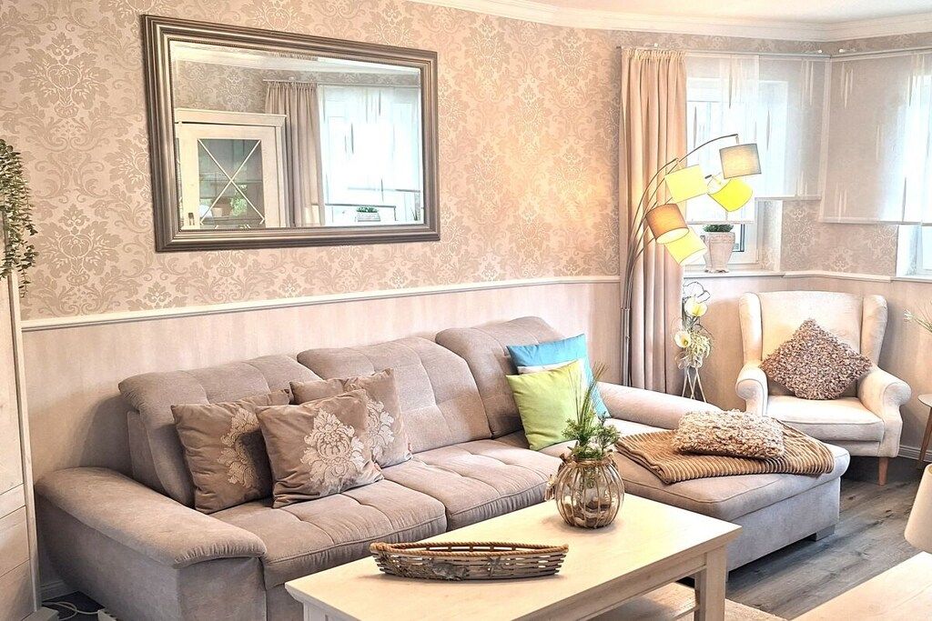 A living room with a sofa, armchair, and a coffee table. There is a mirror on the wall and a lamp on a side table.