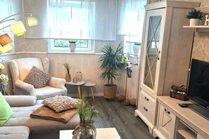A living room with a sofa, armchair, and a television on a cabinet. There are potted plants and a small table with a lamp. - Holiday apartment with 1 bedroom (56332 Burgen)