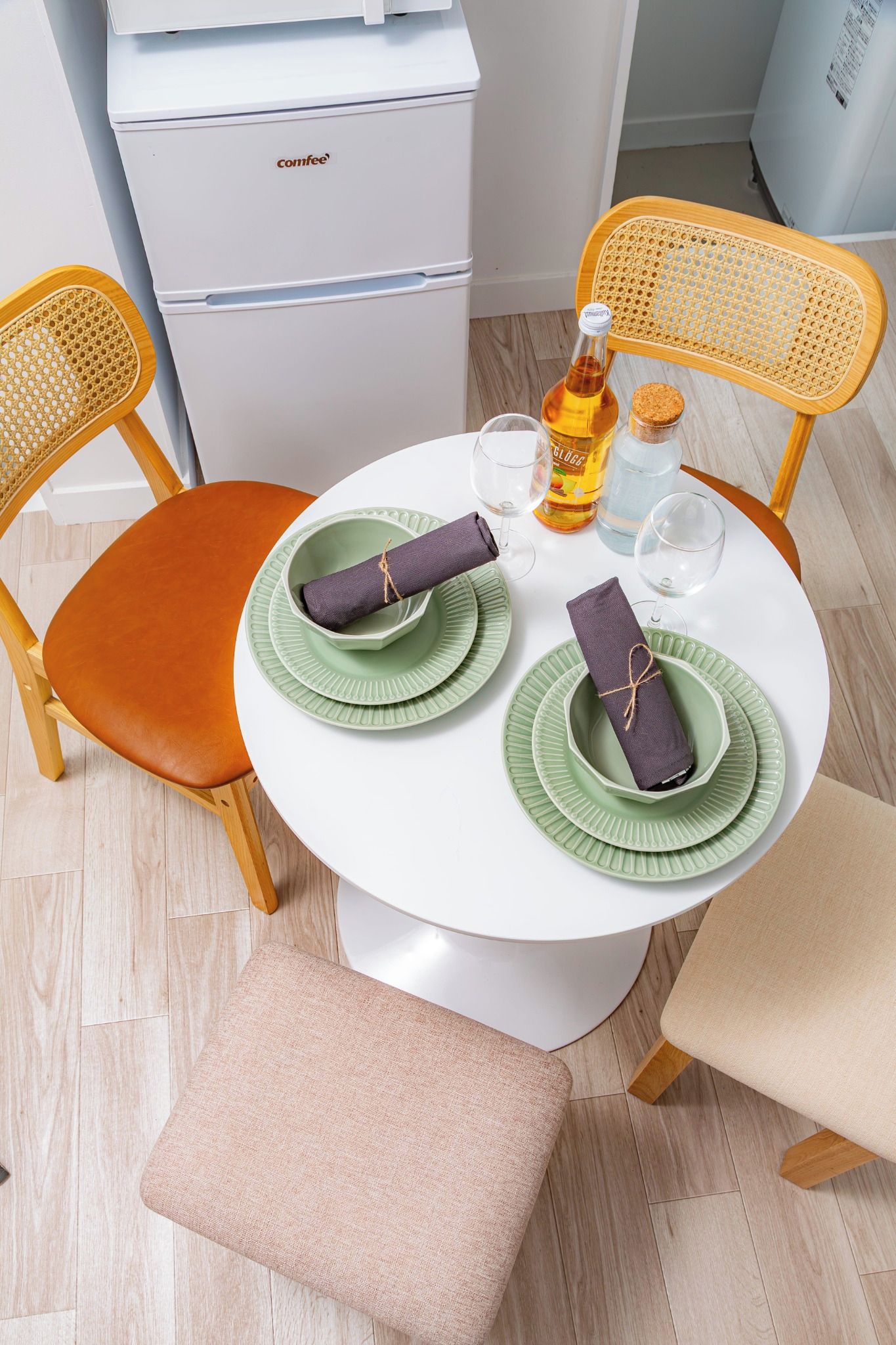 A compact dining area with a white table, two wicker chairs, a bench, and a compact refrigerator.