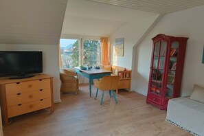 Una habitación con una televisión sobre un mueble de madera, una mesa de comedor con sillas, una librería roja y una ventana con cortinas.