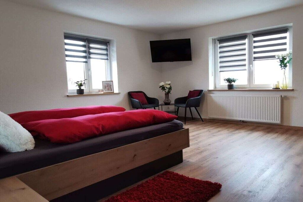 Un dormitorio con una cama, televisión, dos ventanas con persianas, un radiador y una pequeña mesa con sillas.