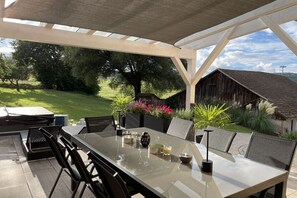 Een terras buiten met een tafel met glazen blad, stoelen en een pergola.