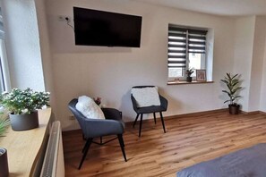 Une pièce dotée d’un téléviseur à écran plat fixé au mur, de deux fauteuils avec des coussins blancs, d’une fenêtre avec des stores et d’une plante en pot.
