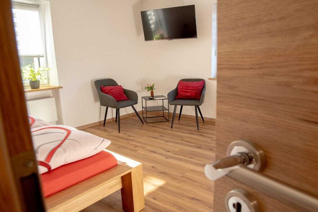 Una habitación con una cama, dos sillones, una mesita, un televisor en la pared y una ventana con una planta.