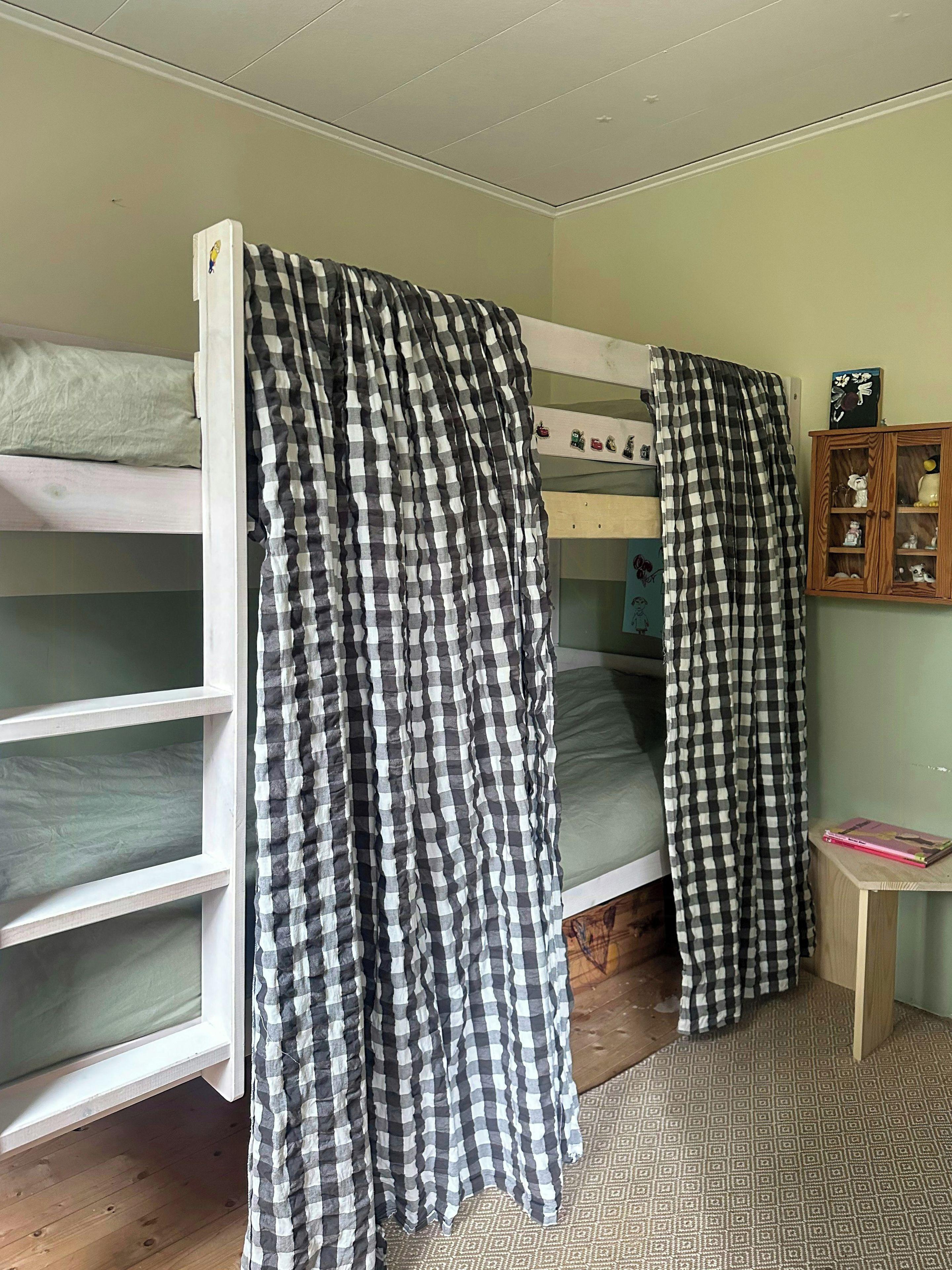 Una camera da letto con un letto a castello, tende a quadretti, un piccolo armadietto in legno con oggetti decorativi e un tappeto a motivi.
