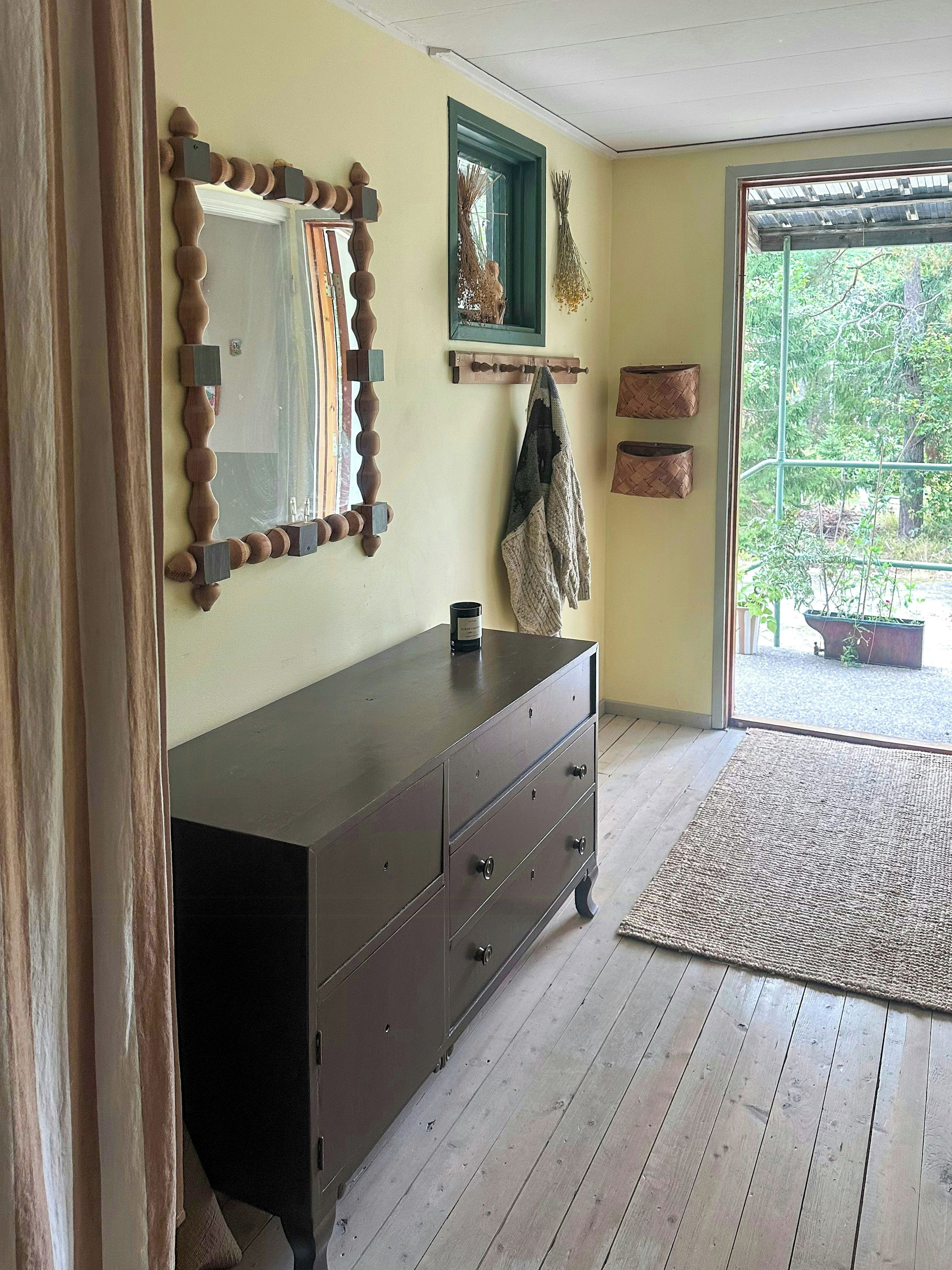 Una habitación con un aparador de madera, un espejo con marco de madera, una ventana con vistas a la vegetación y una puerta que da al exterior.