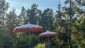 Um pátio externo com mesa e cadeiras, cercado por árvores e dois guarda-sóis.