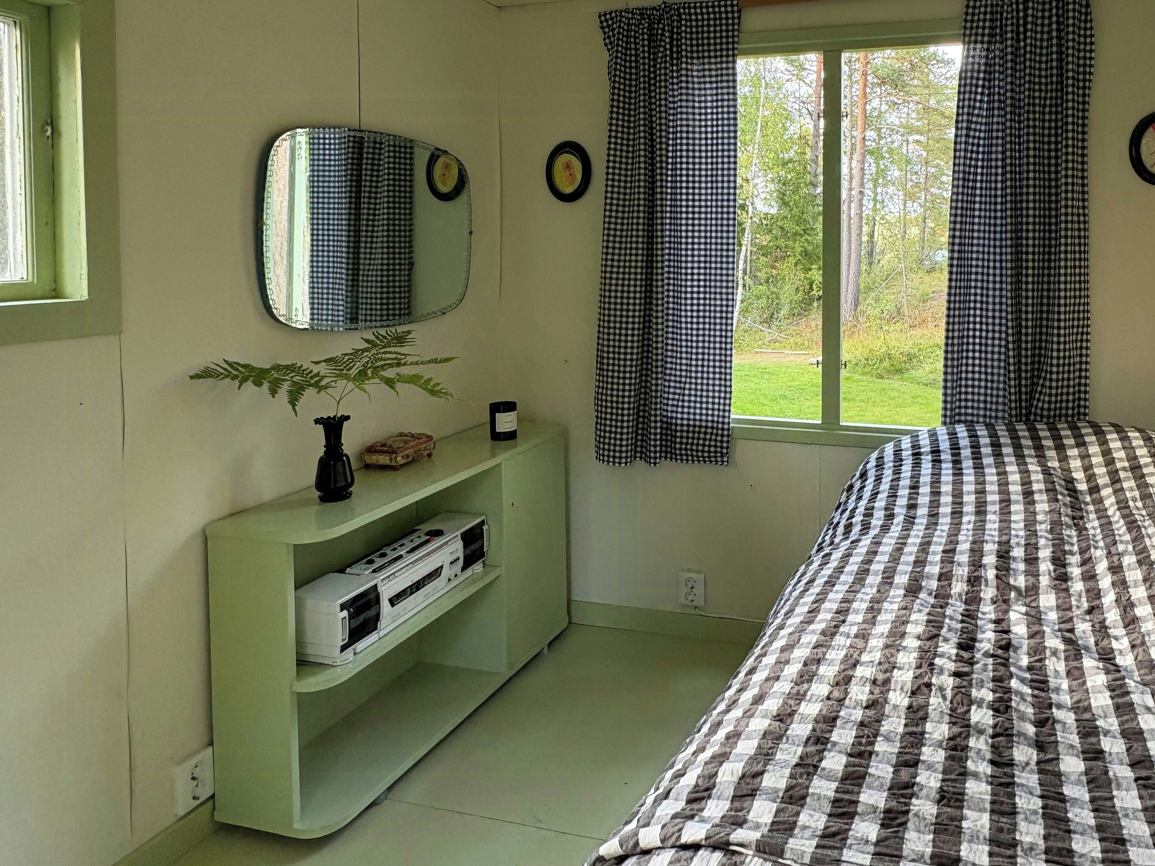 Ein Schlafzimmer mit einem Bett, einer Kommode, einem Spiegel, einem Fenster mit Vorhängen und einer Pflanze.