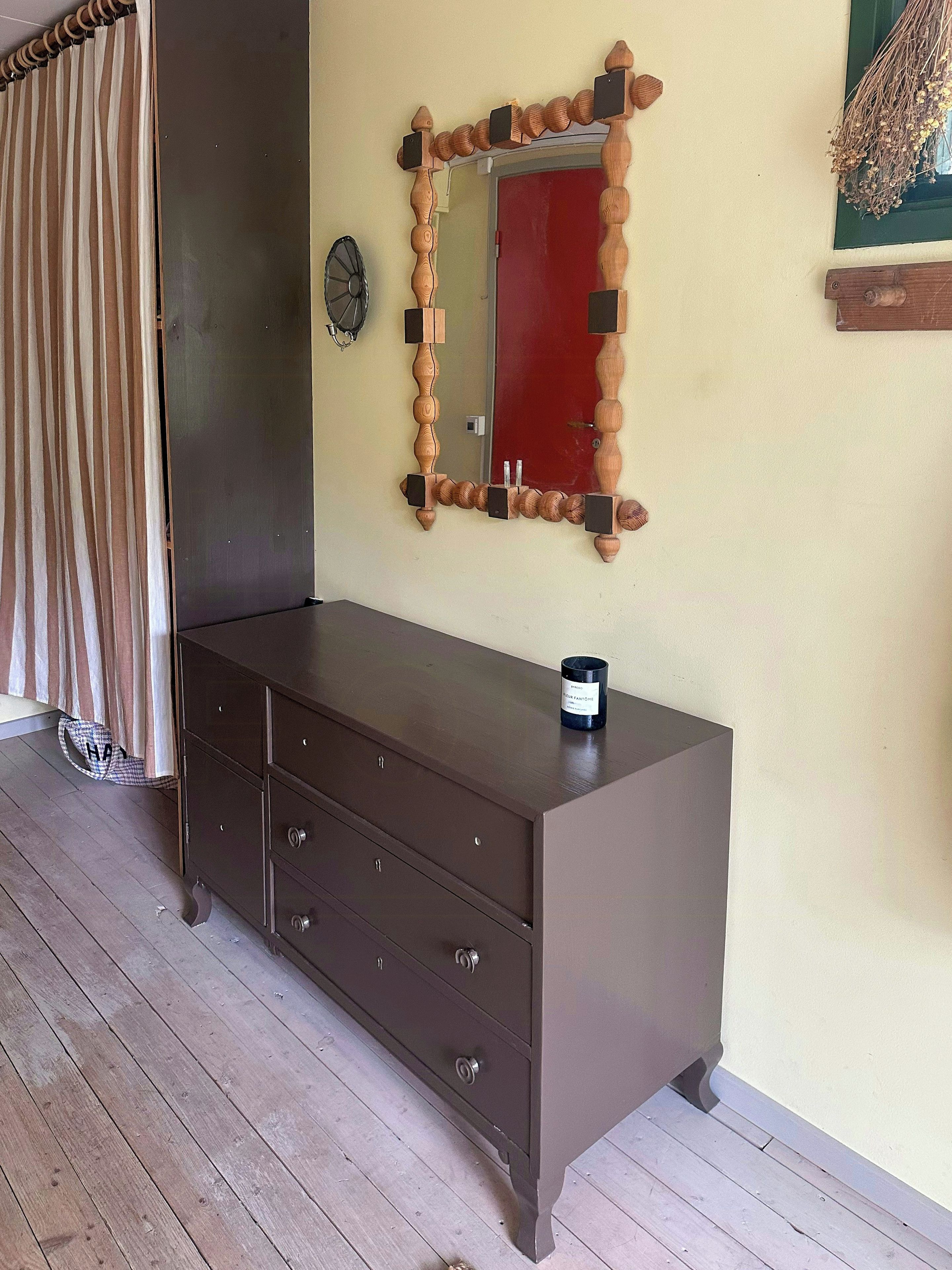 A room with a wooden dresser, a mirror with a unique frame, a red door, and a window with a view.