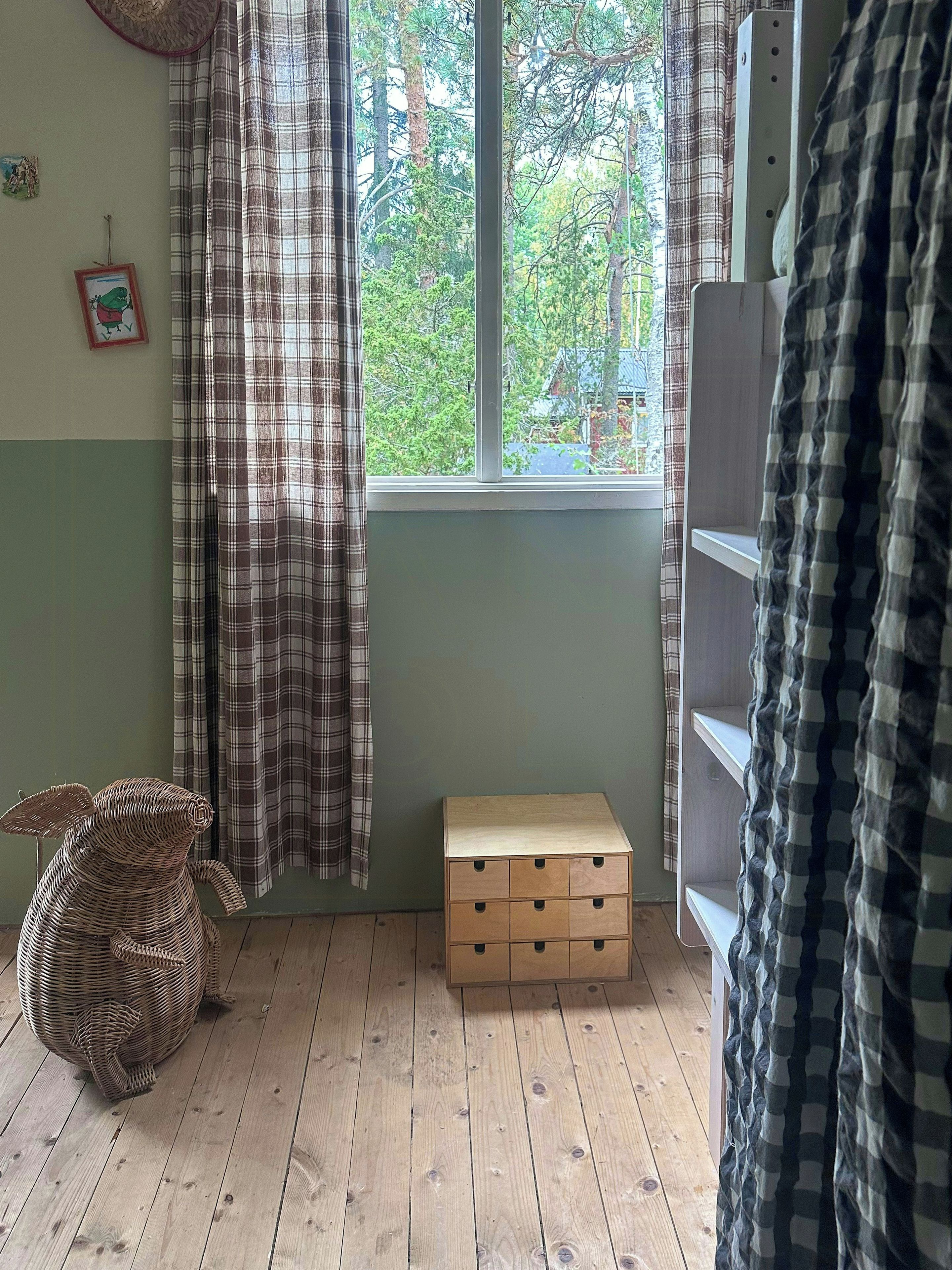 Ett rum med ett fönster, ett trägolv, en liten trälåda, en korg av rotting och en randig gardin.