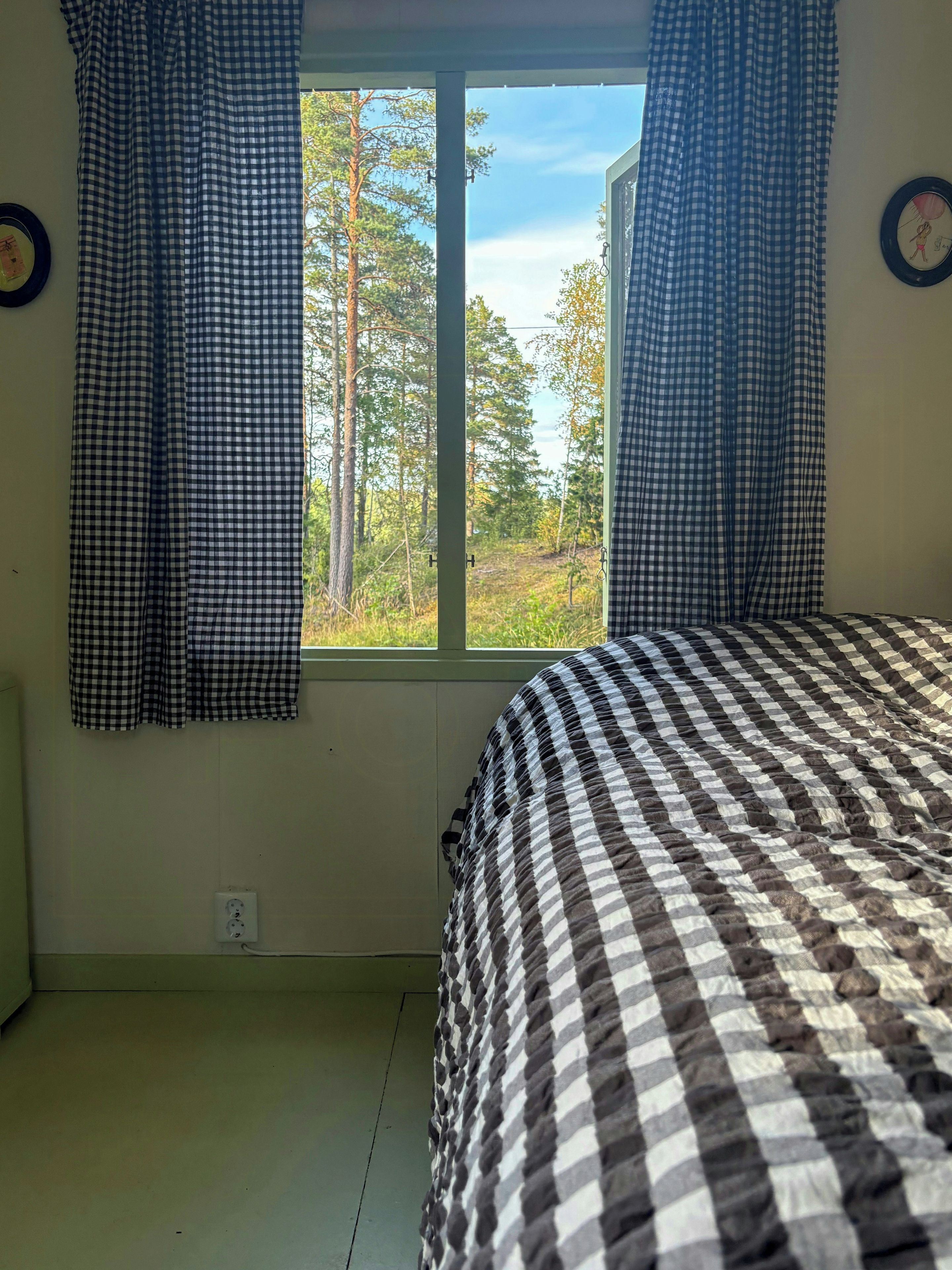 Een slaapkamer met een geruit beddegoed, een raam met gordijnen en uitzicht op bomen.