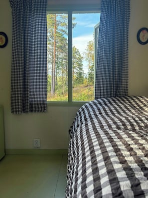Une chambre avec une couvre-lit à carreaux, une fenêtre avec des rideaux et une vue sur les arbres à l’extérieur.