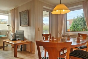 A dining area with a wooden table and chairs, a computer on a desk, and a window with curtains. - Holiday apartment with 1 bedroom (Fehmarn)