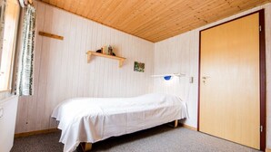 A single-bed room with a wooden ceiling, a small shelf with books, a door, and a window with curtains. - Nydamsgaard "hus lille rede" (Faaborg \/ Bojden)