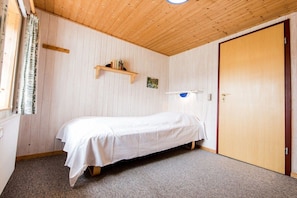 A single-bed room with a wooden ceiling, a small shelf with books, a door, and a window with curtains. - Nydamsgaard "hus lille rede" (Faaborg \/ Bojden)