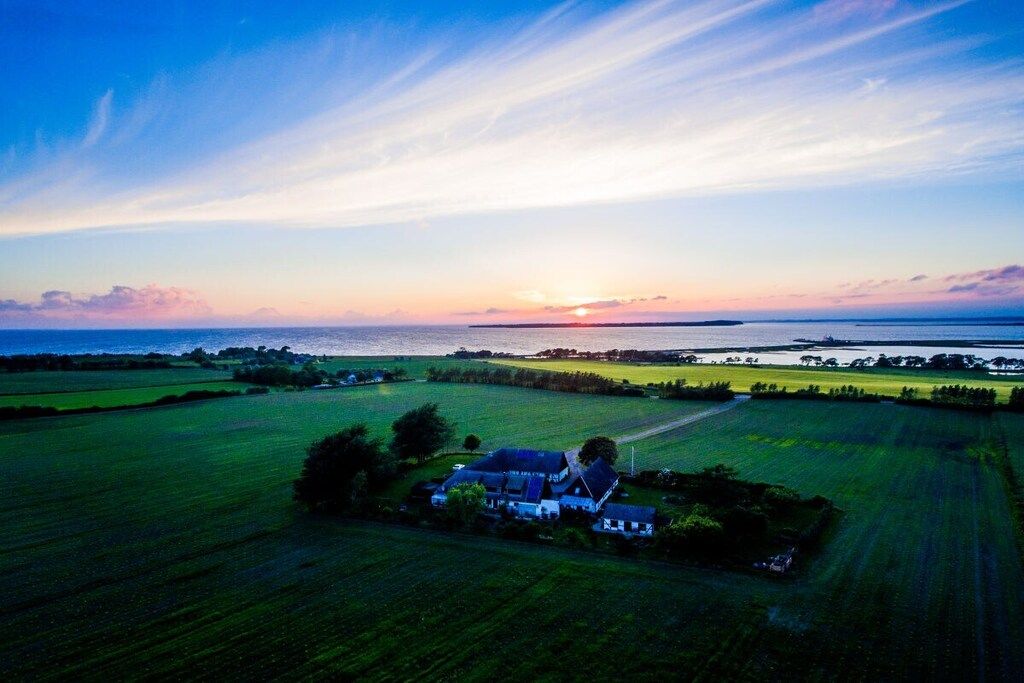 A rural landscape with a house surrounded by fields and a sunset over the water.